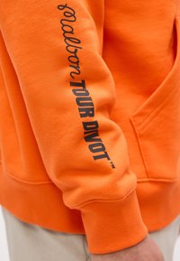 Orange sweatshirt sleeve with black text reading "ballon TOUR DIVOT™" worn over light-colored pants.