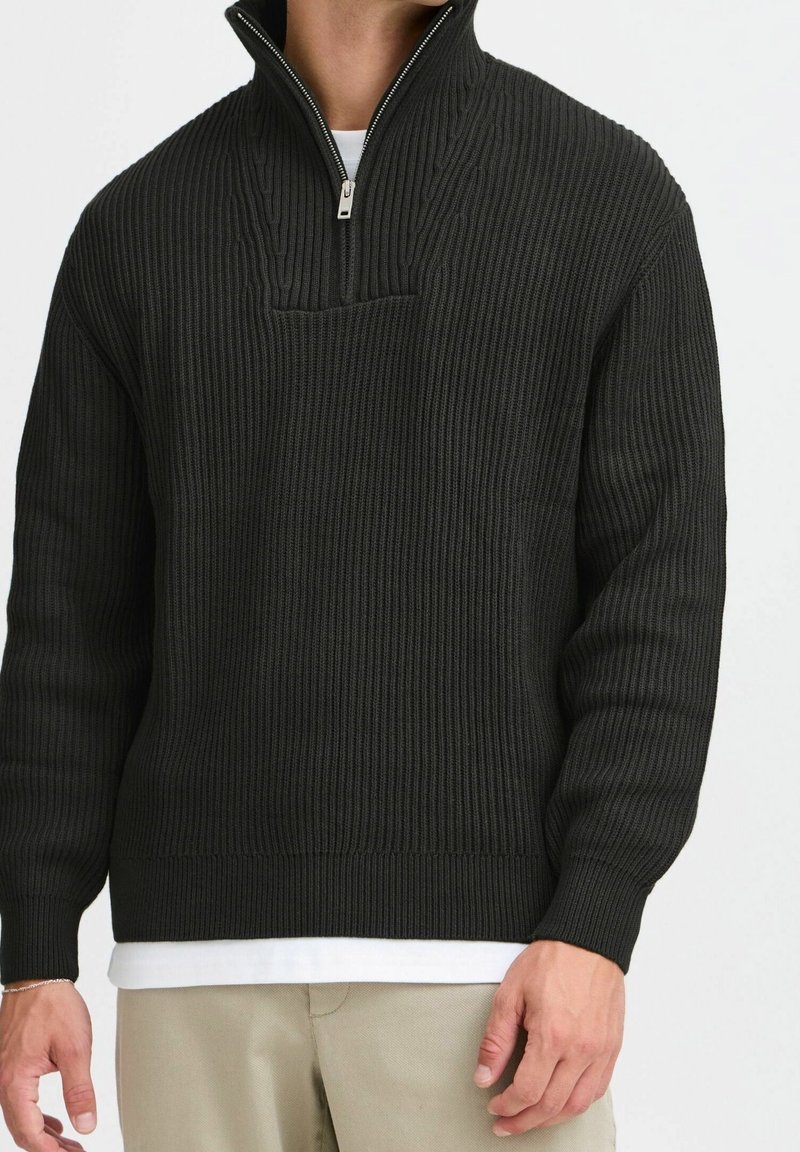 Homme portant un pull à demi-fermeture éclair en maille côtelée noire sur une chemise blanche avec un pantalon beige, debout devant un fond clair uni.