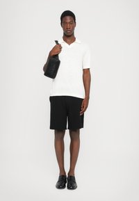 Weißes, strukturiertes Poloshirt, schwarze Shorts und schwarze Slipper. Das Model hält eine schwarze rechteckige Tasche mit geprägtem Text. Einfarbiger Hintergrund.