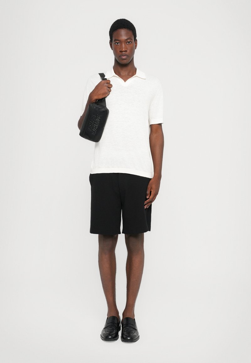 Weißes, strukturiertes Poloshirt, schwarze Shorts und schwarze Slipper. Das Model hält eine schwarze rechteckige Tasche mit geprägtem Text. Einfarbiger Hintergrund.