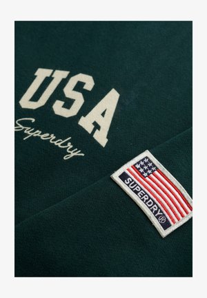Sudadera verde oscuro con el texto en blanco "USA", de la marca "Superdry", y un parche de tela que presenta la bandera de EE.UU. y el logotipo de la marca.