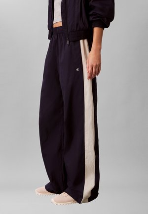 Personne portant un pantalon noir ample avec des bandes blanches sur les côtés, un top court blanc, une veste noire et des baskets beiges, debout sur un fond gris.
