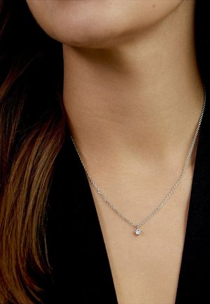 Nahaufnahme des Halses einer Person, die eine filigrane silberne Halskette mit einem kleinen runden Diamantanhänger über schwarzer Kleidung trägt.