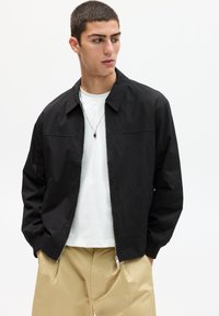Schwarze Bomberjacke mit einem Reißverschluss vorne, umgelegtem Kragen und elastischen Bündchen, getragen über einem weißen T-Shirt und beigen Hosen. Einfaches Design.