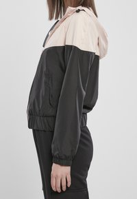 Färgblockerad vindjacka med en ljusrosa övre del och svart nedre sektion, med huva, elastiska ärmmuddar och sidfickor.