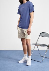 Light blue T-shirt with small Nike logo, beige shorts, white sneakers, and white athletic socks. Background includes a metallic chair and blue floor.