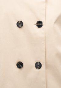 Μπεζ ύφασμα με τέσσερα σκούρα, στρογγυλά κουμπιά τοποθετημένα σε δύο κάθετες σειρές, που εμφανίζουν τρύπες κουμπιών και ραφές λεπτομερειών.