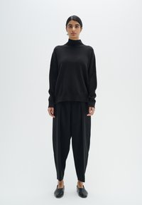 Pulover oversized negru cu guler înalt, purtat cu pantaloni negri largi și pantofi negri fără toc. Design simplu și textură de țesătură netedă.
