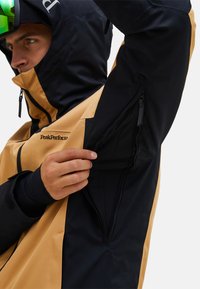 Giacca impermeabile nero e marrone con cappuccio, dotata di tasche con cerniera e finitura texturizzata. Polsini regolabili e marchio ben visibile.