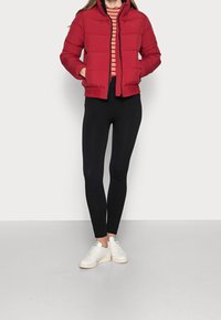 Giacca a vento rossa con design trapuntato, zip frontale e colletto alto, abbinata a leggings neri a coste e sneakers bianche. Top a righe visibile.