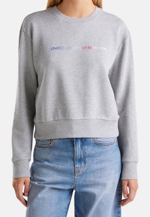 Person iført en grå sweatshirt med flerfarvet "United Colors of Benetton" tekst og blå jeans, stående foran en ensfarvet baggrund.