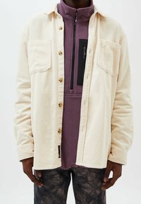 Crème gekleurde corduroy shirtjas met knoopsluiting en twee borstzakken; gedragen over een paarse fleece top met een ritszak.