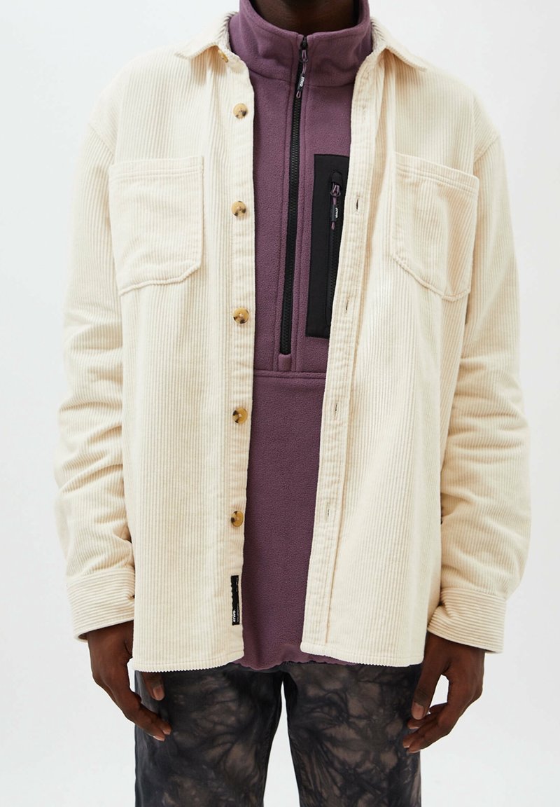 Crème gekleurde corduroy shirtjas met knoopsluiting en twee borstzakken; gedragen over een paarse fleece top met een ritszak.