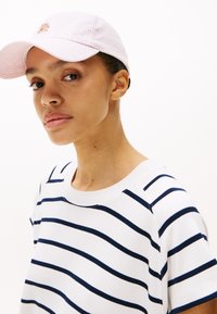 Stripete t-skjorte i hvit med marineblå horisontale striper; myk stoff; brukt under en lys rosa stripete caps med et brodert logo.