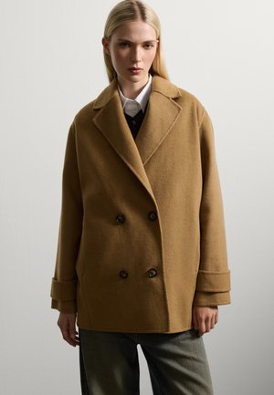 Cappotto in lana marrone doppiopetto con grandi bottoni, revers ampi e polsini larghi; indossato su un gilet nero e una camicia bianca; vestibilità rilassata.