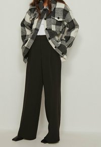 Black and white plaid shirt jacket with two chest pockets, paired with high-waisted, wide-leg dark trousers and pointed black shoes.