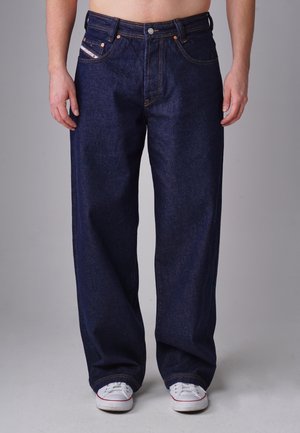 Man wearing loose dark blue jeans and white sneakers standing against a plain gray background.