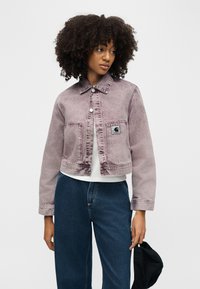 Γυναίκα με σγουρά μαλλιά που φοράει ένα ξεβαμμένο μοβ κοντό τζιν μπουφάν, λευκό πουκάμισο, μπλε τζιν, κρατώντας ένα μαύρο καπέλο στο δεξί της χέρι.