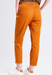 Pantalon chino léger orange à coupe effilée, avec deux poches arrière et ourlets retroussés, associé à des baskets blanches avec des accents bruns.