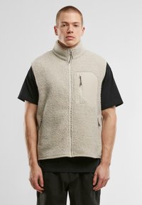 Beige fleece vest with high collar, textured surface, front zip pocket, and side zippers. Worn over a black t-shirt.