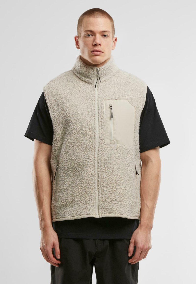 Beige fleece vest with high collar, textured surface, front zip pocket, and side zippers. Worn over a black t-shirt.