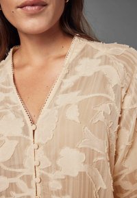 Gennemsigtig, lys beige bluse med blomsterbroderi, V-udskæring, delikate bølgede kanter og stofbetrukne knapper foran.