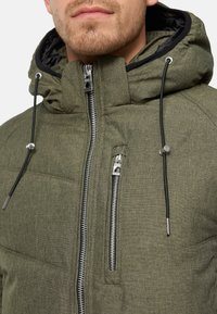 Olijfgroene pufferjack met een hoge kraag, ritsafsluitingen en verstelbare koordjes. Heeft een textuur, zachte stof en contrasterende voering.