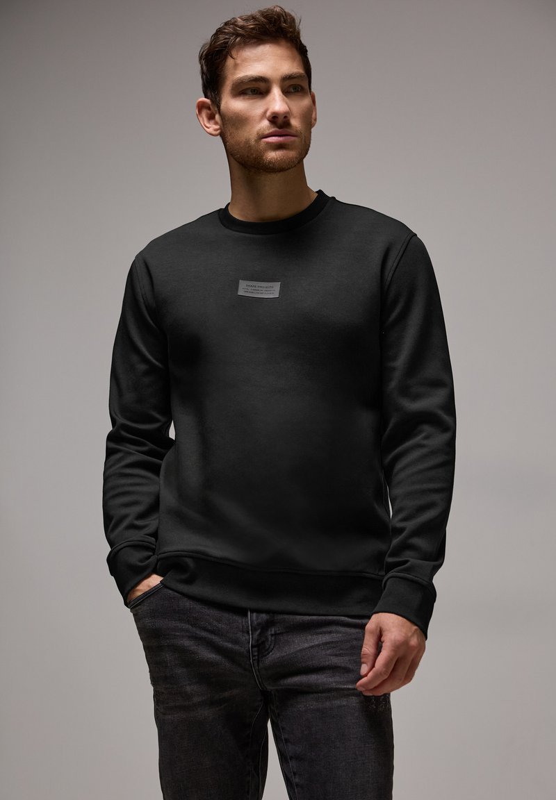 Sort sweatshirt i blød bomuldsblanding, med rund hals og lange ærmer, samt et lille mærkelogo på brystet. Bæres med mørke jeans.