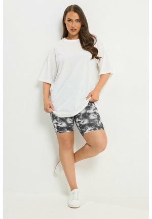 Frau trägt übergroßes weißes T-Shirt und schwarz-weiße Biker-Shorts im Batik-Look, steht mit einem angewinkelten Bein vor einer weißen Wand.