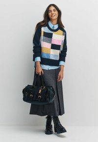 Kobieta w wielokolorowym swetrze z naszywkami, niebieskiej koszuli, długiej szarej spódnicy i czarnych wojskowych butach, trzymająca dużą czarną torebkę na jednolitym tle.