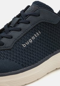 Tengerészeti sportcipő szőtt textil felsőrésszel, sima bőr részletekkel és krémszínű gumitalppal. Az oldalon "bugatti" logó található.
