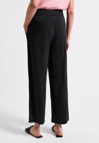 Black, loose-fitting trousers with an elastic waistband, featuring two back pockets and a smooth texture. Worn with flat sandals.