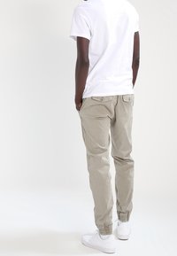 Beige cargo-byxor med elastiska muddar, två bakfickor och en avslappnad passform, kombinerat med en vit t-shirt med kort ärm och vita sneakers.