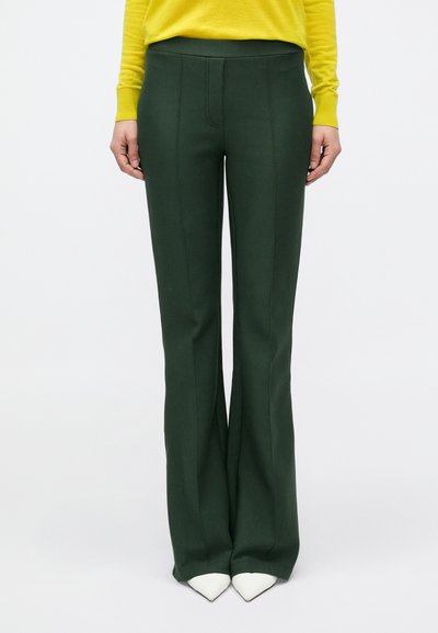 Pantalon évasé vert foncé avec une texture lisse, taille haute et détails de coutures verticales. Associé à des talons blancs pointus au bas.