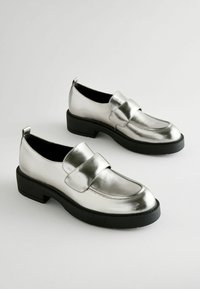 Mocasines metálicos plateados con un acabado suave, tacón negro apilado y una correa decorativa en la parte delantera. Diseño de suela plana.