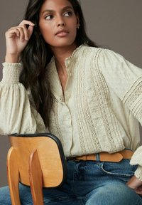 Chemisier couleur crème en tissu texturé, haut col froncé, détails en dentelle intricats et manches volumineuses. Porté avec une ceinture beige et un jean bleu.