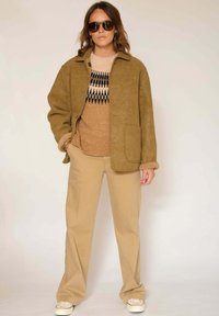 Veste en daim beige avec une poche, portée par-dessus un pull beige à motifs. Assortie à un pantalon beige et des baskets blanches. Lunettes de soleil foncées portées.