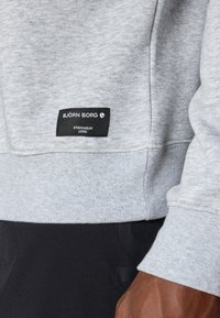 Sweatshirt cinza com um tecido suave e texturizado, apresentando um rótulo preto com texto branco que detalha a marca e a origem. Corte casual com punhos canelados.
