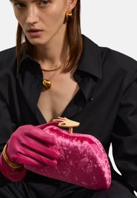 Clutch in velluto rosa con una chiusura a forma di mano dorata, tenuta da una persona che indossa guanti rosa. Visibile una camicia nera e gioielli dorati.