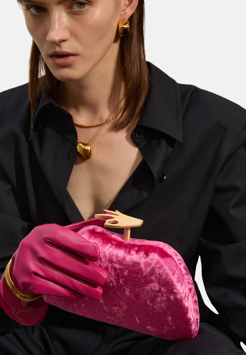 Clutch in velluto rosa con una chiusura a forma di mano dorata, tenuta da una persona che indossa guanti rosa. Visibile una camicia nera e gioielli dorati.