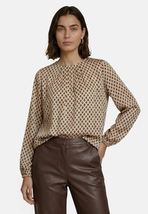 Femme aux cheveux bruns portant un chemisier beige à motifs manches longues, rentré dans un pantalon en cuir marron foncé taille haute, la main dans la poche.