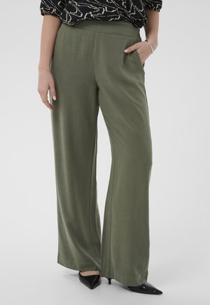 Frau trägt olivgrüne, weit geschnittene Hosen und schwarze spitze High Heels, Hand in der Tasche, steht vor einem schlichten Hintergrund.