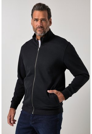 Mand med skæg iført sort lynlåstrøje og blå jeans, poserer med den ene hånd i jakkelommen mod en ensfarvet baggrund.