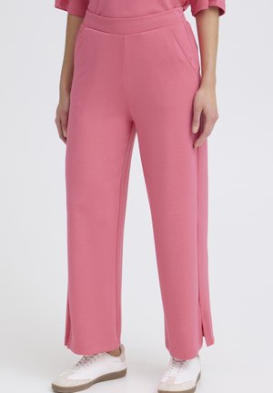 Personne portant un pantalon ample rose avec des poches latérales et de petites fentes sur les côtés, associé à des baskets blanches sur un fond uni.