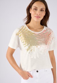 Lichtbeige korte mouw t-shirt met gouden en koraalkleurige bladontwerpen met metallic accenten. De stof heeft een subtiele textuur.
