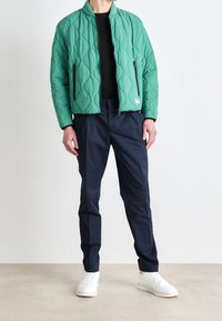 Quiltet grøn jakke med ståkrave, lynlåslommer og navy bukser. Modellen har også hvide sneakers på.