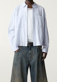 Jasnoniebieska, teksturowana koszula z guzikami z dwoma kieszeniami na piersi, założona na biały top na ramiączkach, w zestawieniu z luźnymi, wyblakłymi szortami dżinsowymi.