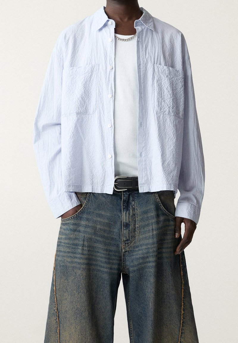 Jasnoniebieska, teksturowana koszula z guzikami z dwoma kieszeniami na piersi, założona na biały top na ramiączkach, w zestawieniu z luźnymi, wyblakłymi szortami dżinsowymi.
