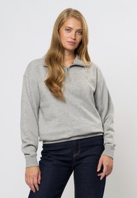 Sivá mikina s zipsovým golierom, vyrobená z mäkkej látky. Obsahuje rebrované manžety a diskrétne logo na hrudi, kombinovaná s tmavými džínsami.