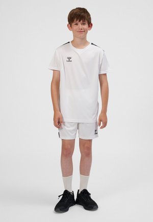 Completo da calcio bianco con maglia a maniche corte e pantaloncini, con dettagli neri, realizzato in tessuto leggero. Indossato con scarpe da ginnastica nere.